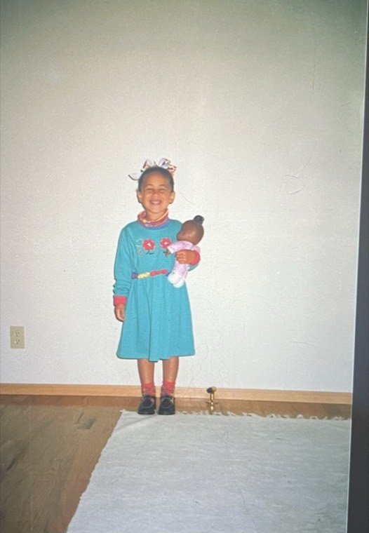 Alexis as a child in a teal dress, alone in an empty room, holding a brown baby doll.