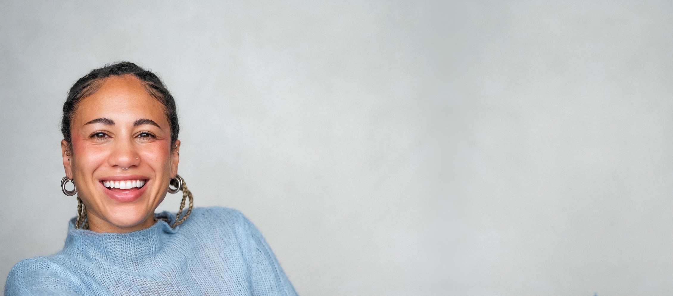 Alexis Kimball seated sideways in a mustard yellow chair, wearing a pale blue mohair sweater, laughing, against a wide grey wall.