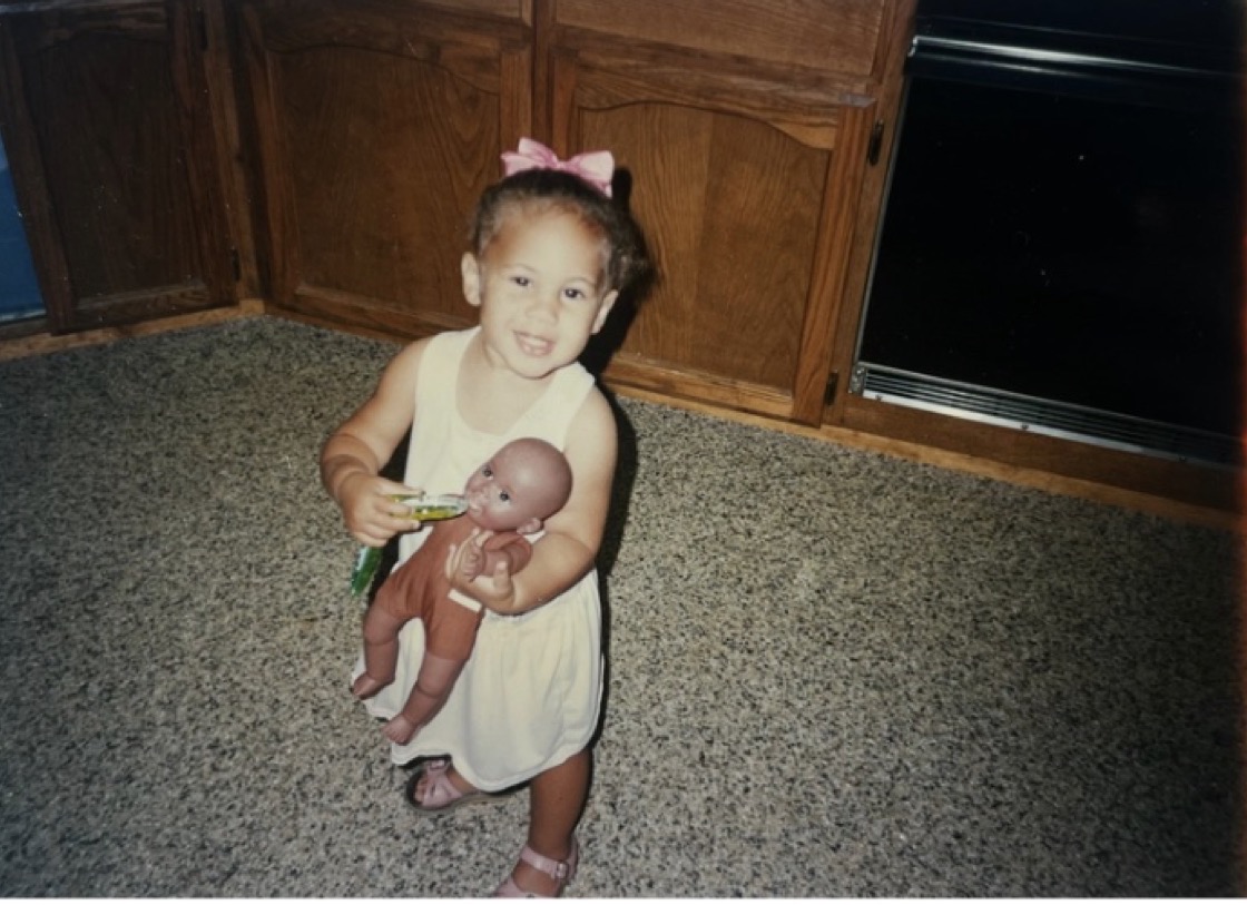 Alexis as a toddler in a white dress with a pink bow, standing in a kitchen holding a brown baby doll.