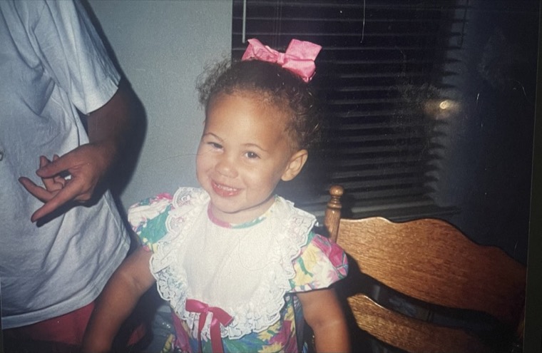 Alexis as a toddler in a lace dress with a pink bow, an adult standing beside her.